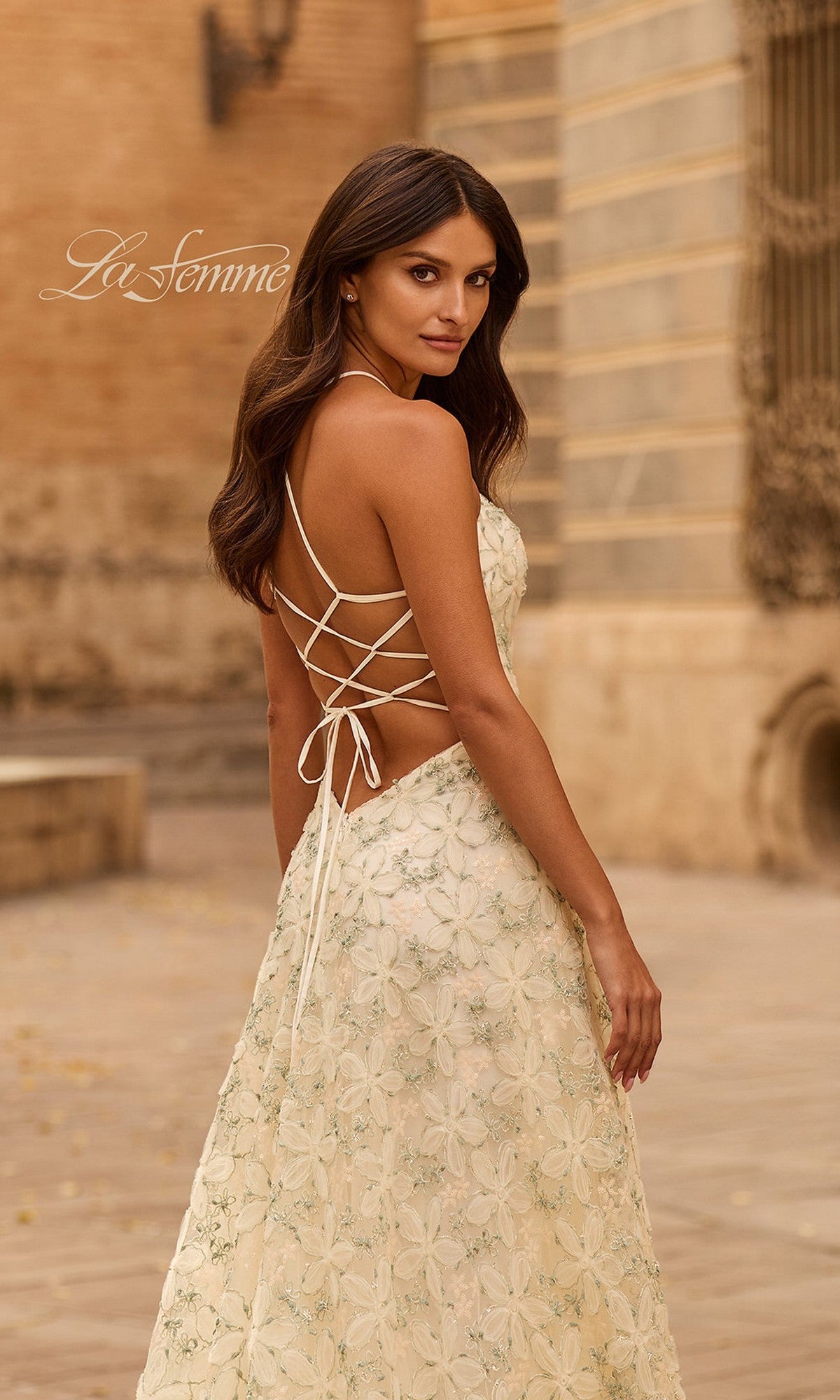 A woman wears the La Femme 33865 cream A-line prom dress with floral lace embroidery and a lace-up open back, standing outdoors and looking over her shoulder.